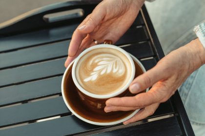 close-up of a latte with intricate art on a wooden table, held by hands.