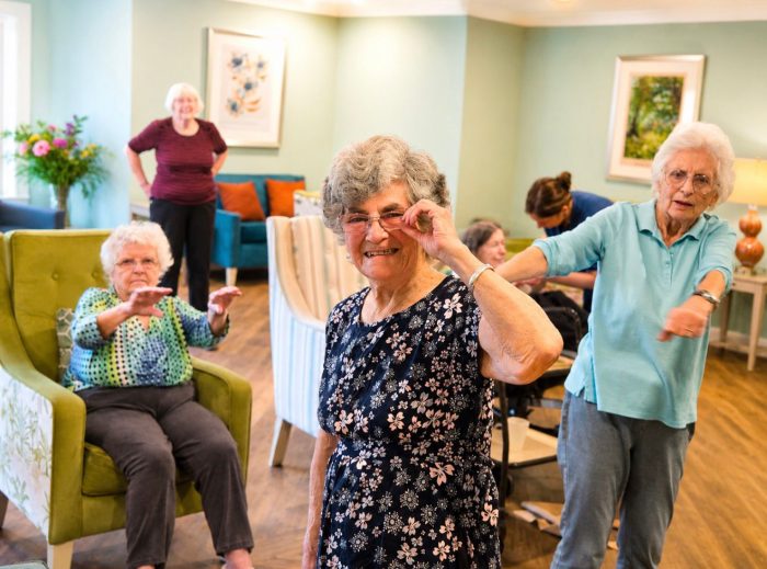84-year-old resident Sylvia Hawley smiling in a floral dress while leading a group chair exercise and dance class for residents at Gateford Lodge Care Home.